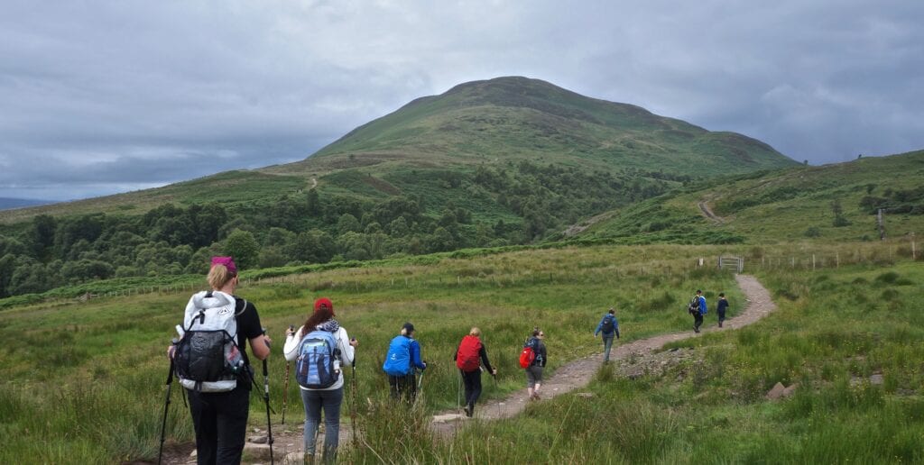 Group hiking on a trail through lush green hills under a cloudy sky.